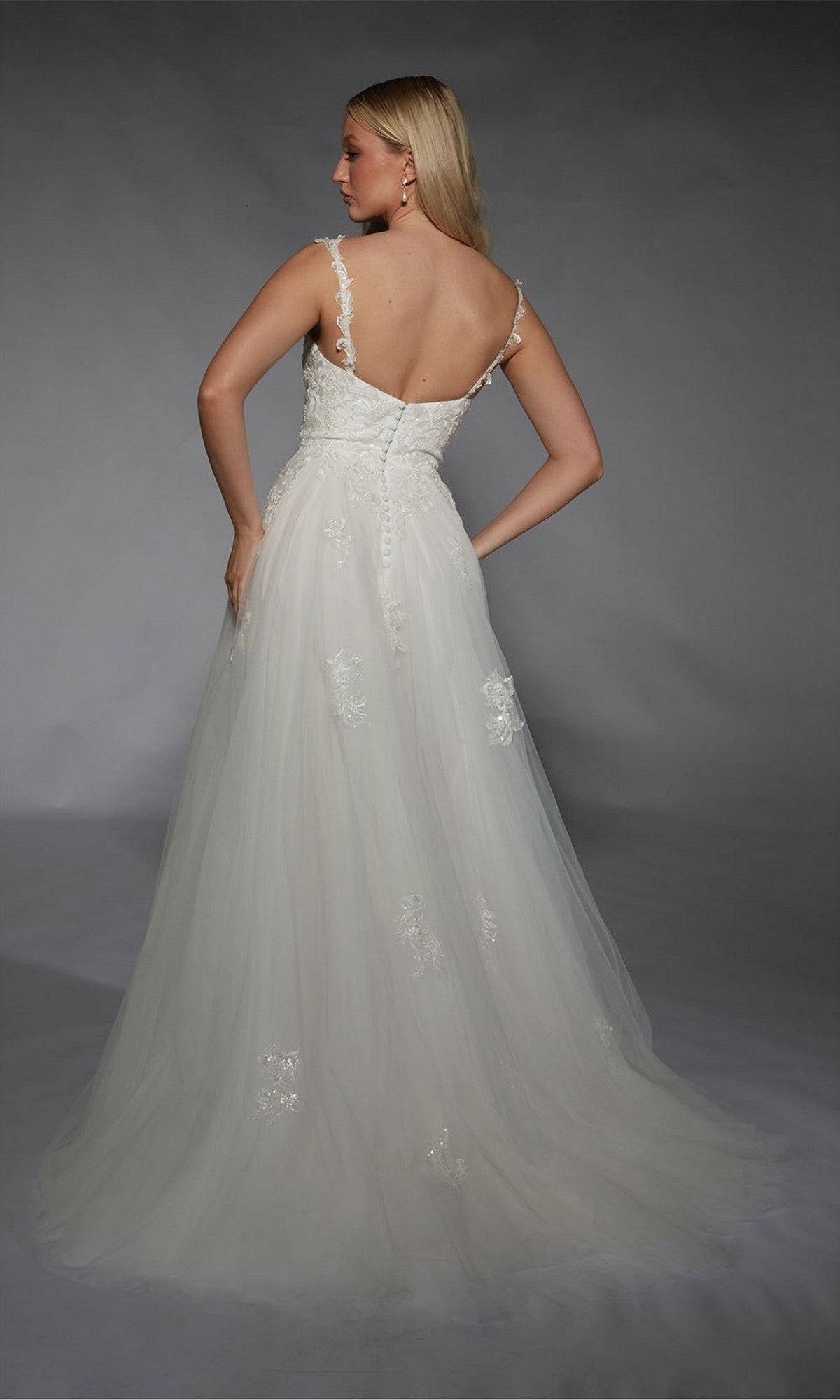 A woman faces away, wearing the Long White Dress: Alyce 7124, featuring delicate lace appliques and a flowing A-line silhouette, against a plain gray background.